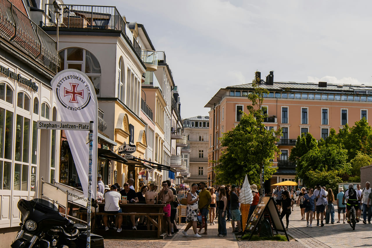 A busy seaside promenade: people at cafe tables and browsing shopfronts, others walking and cycling past an ice cream stand and a menu board, with a street sign naming the square.